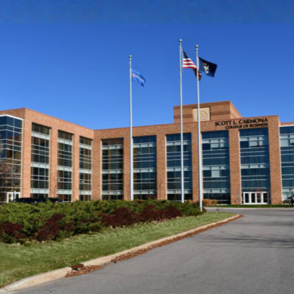 Front exterior of Saginaw Valley State University’s Scott L. Carmona College of Business with brick and glass façade, flags in front, and modern architectural design.