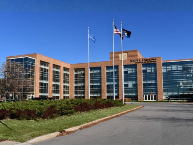 Front exterior of Saginaw Valley State University’s Scott L. Carmona College of Business with brick and glass façade, flags in front, and modern architectural design.